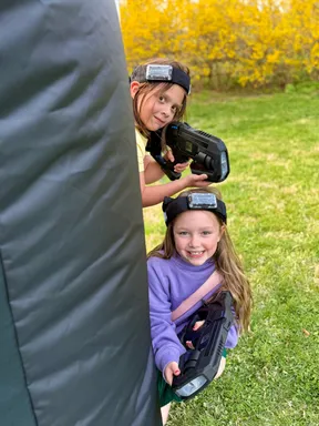 Two girls teaming up behind an inflatable bunker at a backyard mobile laser tag birthday party for kids