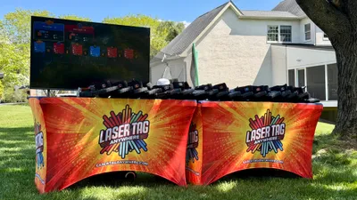 Pro-grade Laser Tag Anywhere equipment table with branded covers, taggers, and a live TV scoreboard at a backyard event